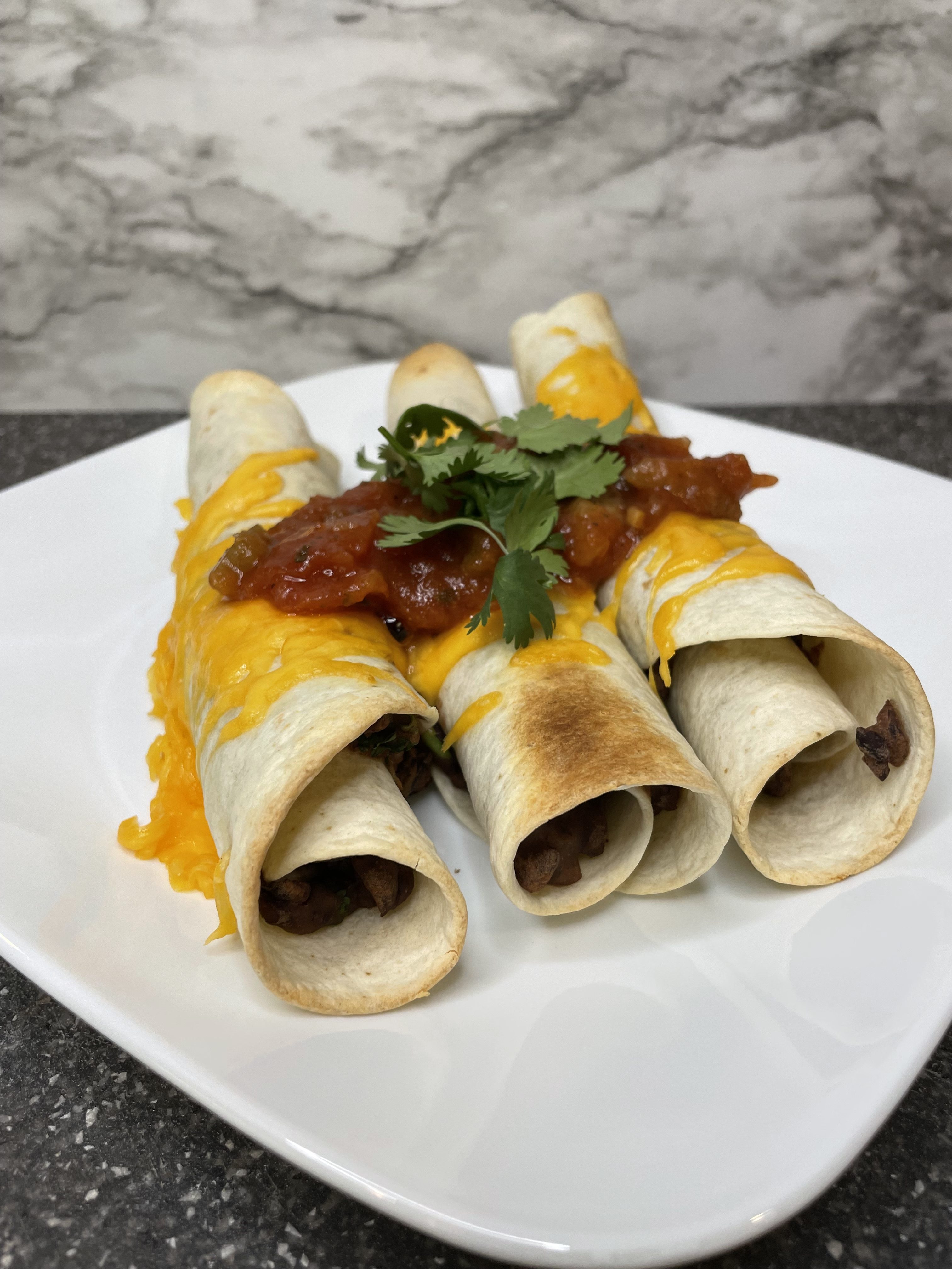 Three black bean taquitos covered in cheese, salsa, and cilantro served on a white plate.